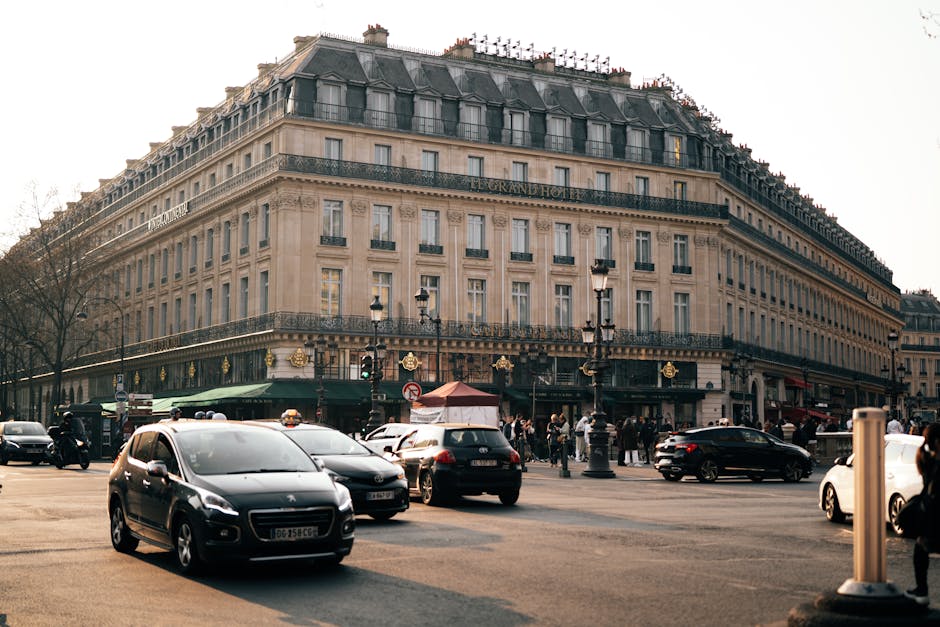 A bustling Parisian street scene with cars and pedestrians in front of a historic hotel facade.