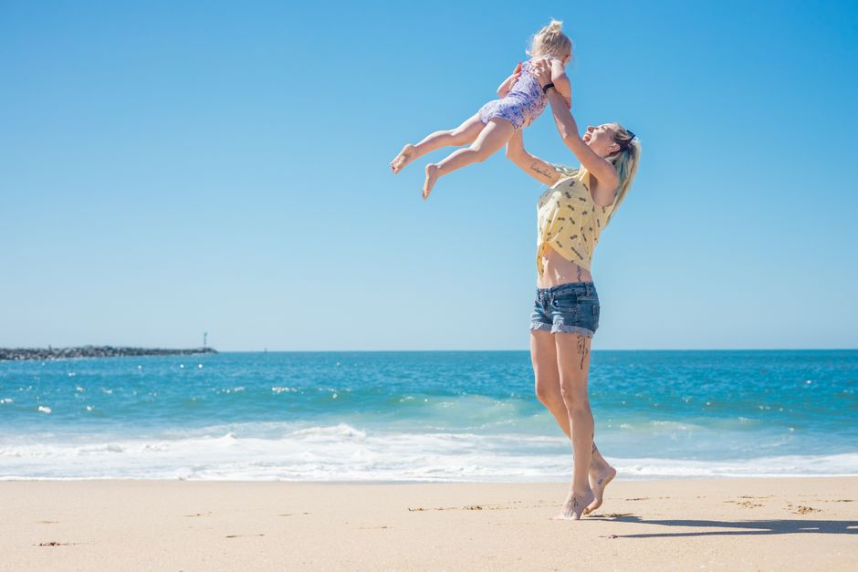 A mother joyfully lifts her daughter in the air at a sunny beach, capturing a moment of family fun.