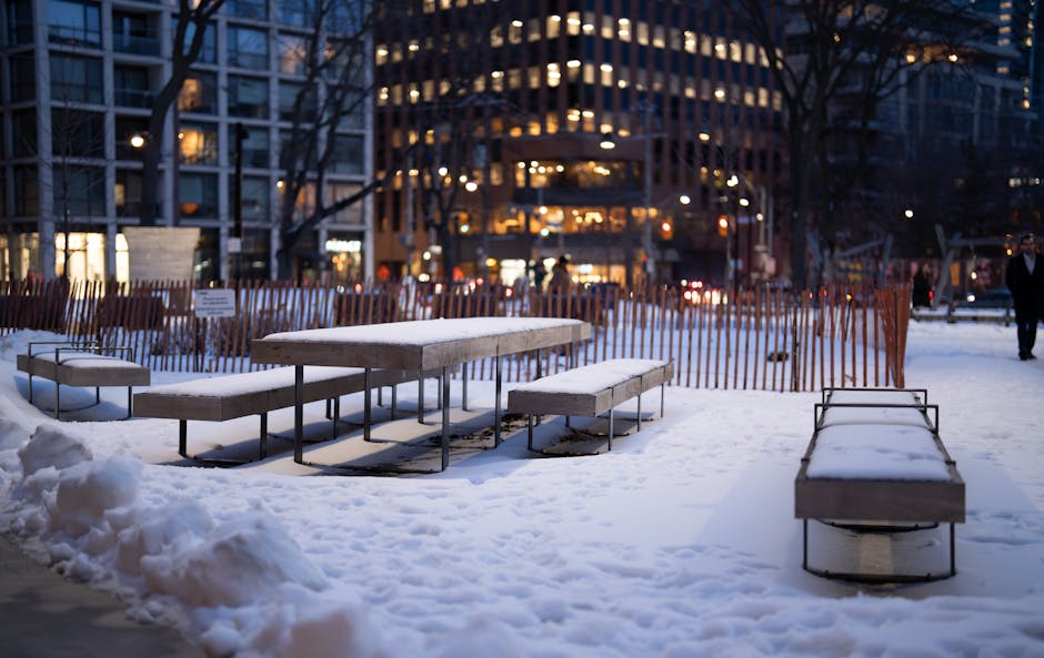 A cozy winter evening scene of a snow-covered urban park in downtown Toronto, showing benches and city lights.
