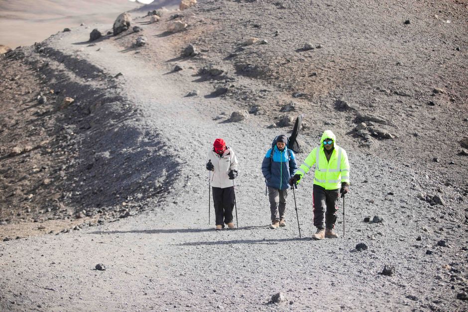 Three hikers traverse a rocky path on a volcanic landscape, equipped with trekking gear.